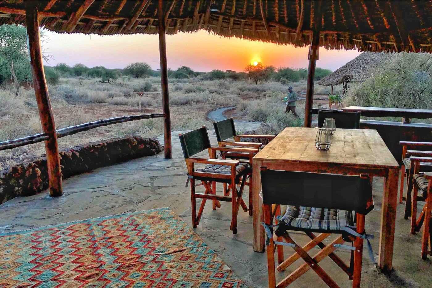 Outdoor dining table set under a thatched roof at sunset at Amboseli Bush Camp.