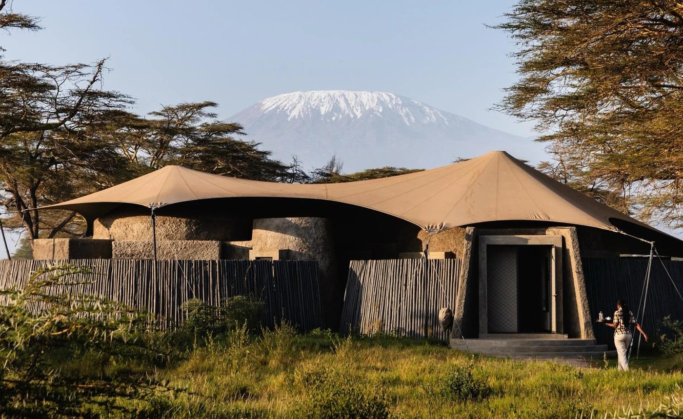 Canvas-roofed suites and lounge structure set among acacia trees below Kilimanjaro at Angama Amboseli.