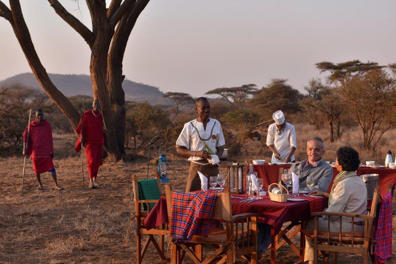 Maasai staff serving an outdoor table set beneath acacia trees at Elerai Camp.