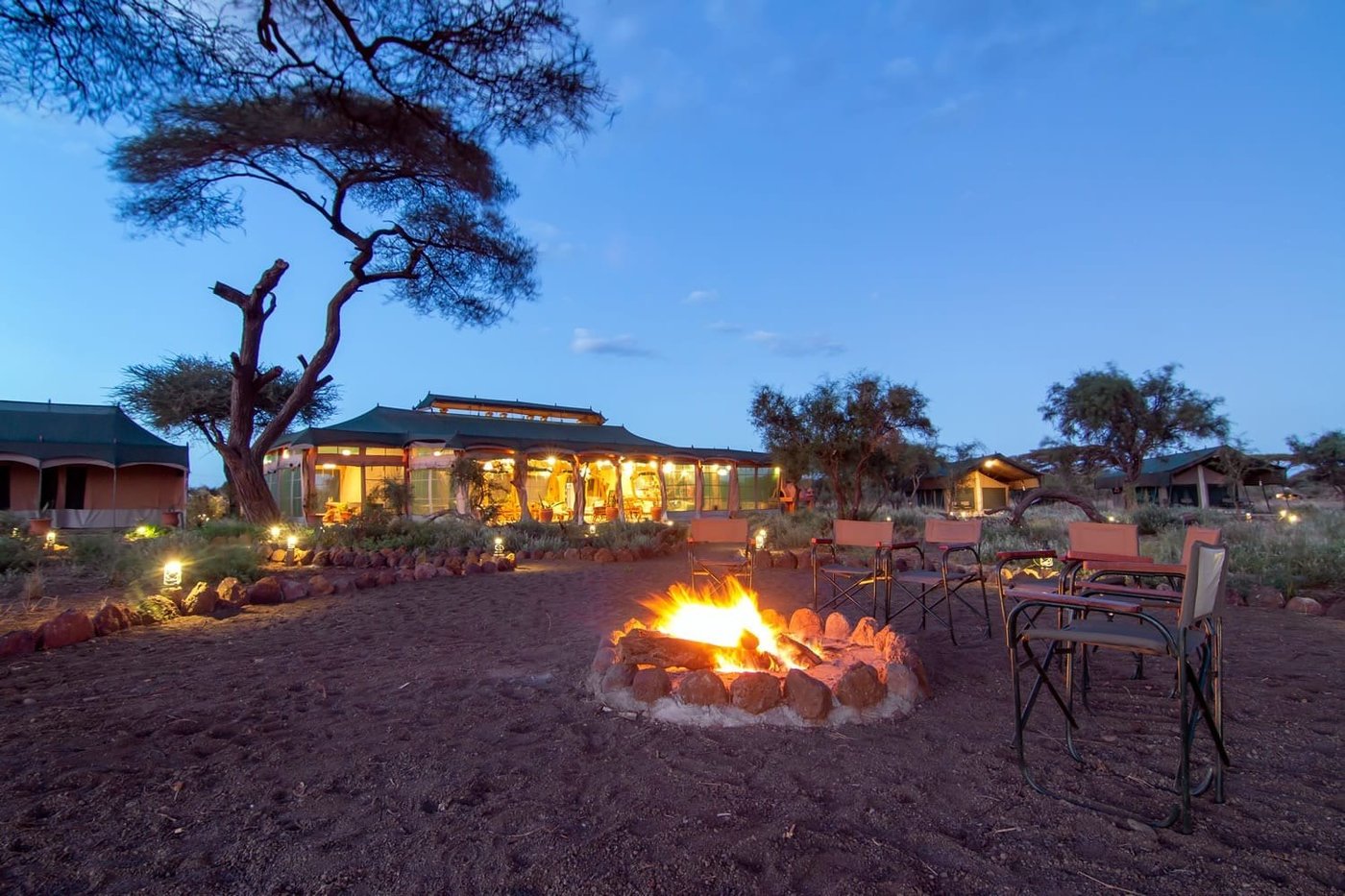 Lit lodge buildings and a glowing campfire under acacia trees at dusk at Kibo Safari Camp, Amboseli.