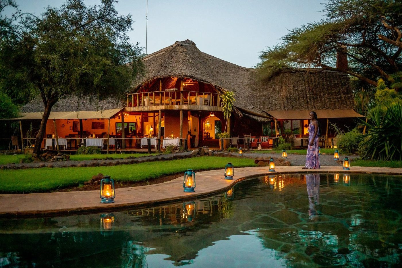 Thatched lodge buildings lit at dusk reflected in the pool at Tawi Lodge, Amboseli.